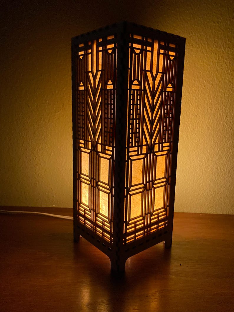 May include: A wooden lamp with a geometric design and a warm, yellow light emanating from the inside. The lamp is on a wooden surface.