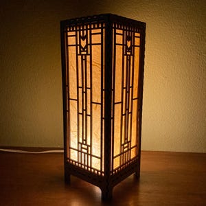 May include: A wooden lamp with a geometric design and a paper shade. The lamp is lit and casting a warm glow.
