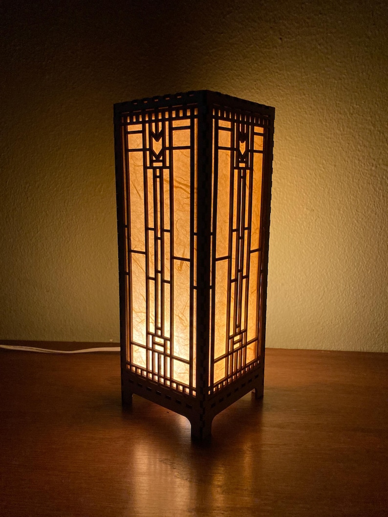 May include: A wooden lamp with a geometric design and a paper shade. The lamp is lit and casting a warm glow.