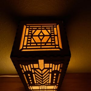 May include: A wooden lamp with a geometric design, lit from within, casting a warm glow. The lamp features a square base and a rectangular shade with intricate cutouts.