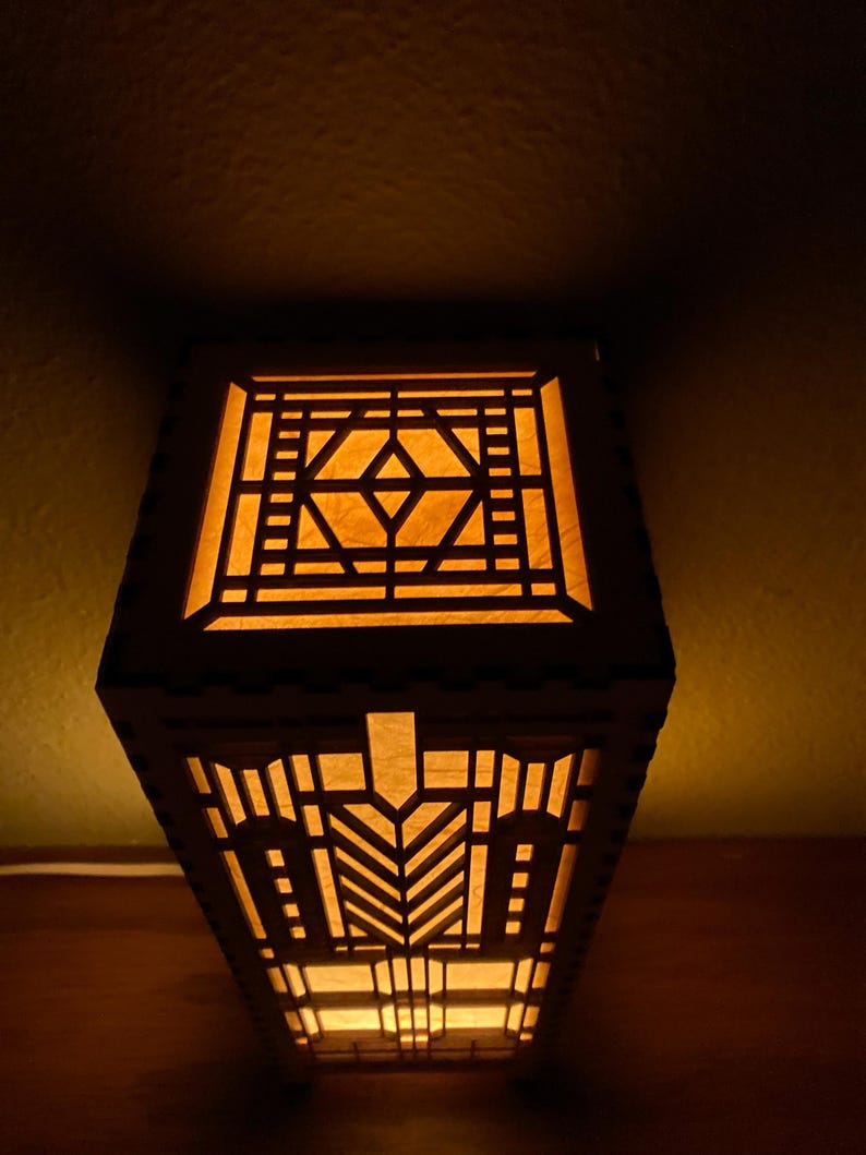 May include: A wooden lamp with a geometric design, lit from within, casting a warm glow. The lamp features a square base and a rectangular shade with intricate cutouts.