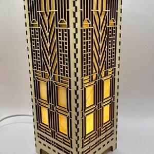 May include: A wooden lamp with a geometric design. The lamp is lit and the light shines through the cutouts in the wood. The lamp has a square base and a rectangular body.