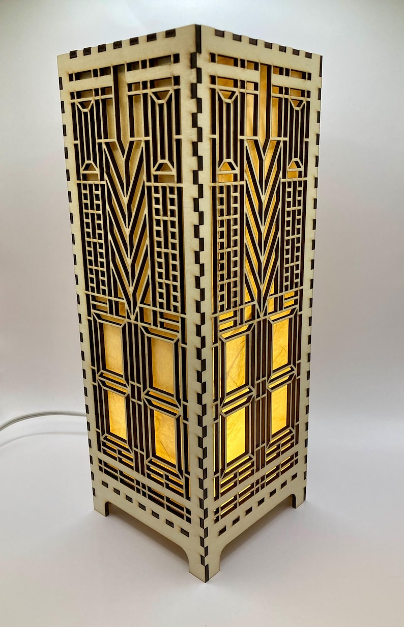 May include: A wooden lamp with a geometric design. The lamp is lit and the light shines through the cutouts in the wood. The lamp has a square base and a rectangular body.