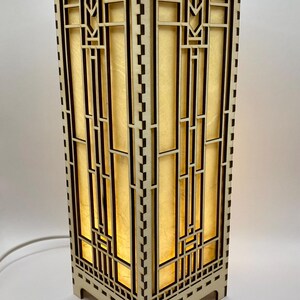 May include: A wooden table lamp with a geometric design. The lamp has a light brown finish and a clear glass shade. The lamp is lit and casting a warm glow.