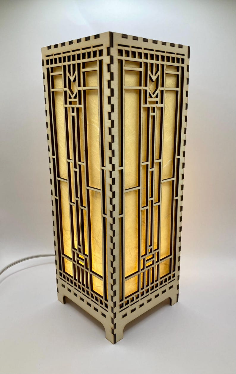 May include: A wooden table lamp with a geometric design. The lamp has a light brown finish and a clear glass shade. The lamp is lit and casting a warm glow.