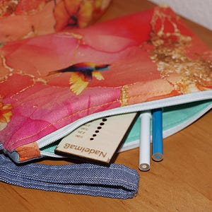 Könnte beinhalten: Eine Reißverschlusstasche mit floralem Druck und einer blauen Denim-Handschlaufe. Die Tasche ist geöffnet und zeigt ein hölzernes Nadelmaß und zwei Bleistifte. Die Innenseite ist helltürkis. Die Tasche hat ein farbenfrohes Blumendesign mit einem Vogel.