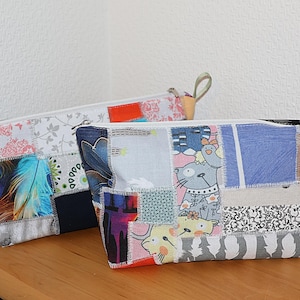 Könnte beinhalten: Zwei Patchwork-Reißverschlusstaschen mit verschiedenen Stoffmustern, darunter Blumen-, Feder- und Cartoon-Katzen-Designs. Die Taschen sind rechteckig mit Reißverschluss und einer kleinen Schlaufe. Sie liegen auf einer Holzoberfläche.