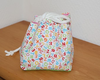 Könnte beinhalten: Ein kleiner Beutel mit Kordelzug aus weißem Stoff, der mit bunten Buchstaben bedeckt ist. Die Buchstaben sind in verschiedenen Farben, darunter Rot, Blau, Gelb und Grün. Der Beutel hat einen weißen Kordelzug und eine rosa Innenverkleidung.