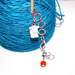Könnte beinhalten: Ein weißer Keramik-Schaf-Anhänger, der an einer silbernen Kette mit roten Perlen befestigt ist. Die Kette ist an einer blauen Garnrolle befestigt.