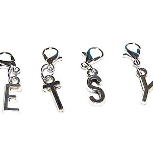 Könnte beinhalten: Vier silberne Buchstaben-Anhänger aus Metall mit Karabinerhaken. Die Buchstaben ergeben "ETSY".