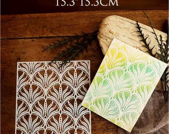 May include: White stencil with a repeating fan-shaped design, next to a watercolor painting with a similar pattern in yellow and green. The stencil measures 15.3cm x 15.3cm. The background is a dark wood surface.