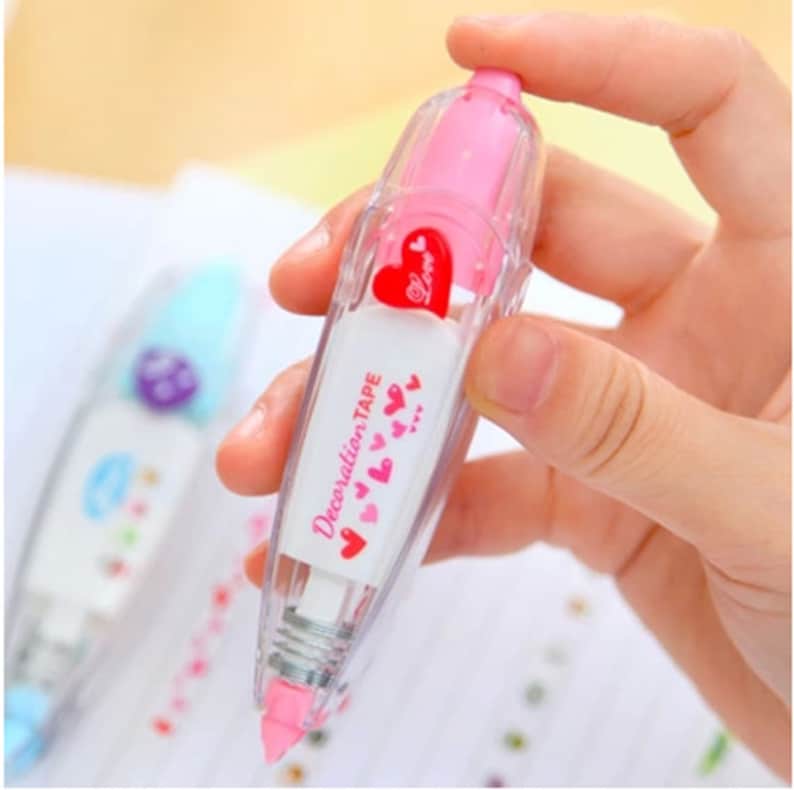 K&ouml;nnte beinhalten: Ein durchsichtiger Plastikspender mit rosa Deckel und einem wei&szlig;en Etikett mit der Aufschrift "Decoration Tape" mit Herzen und dem Wort "Love".