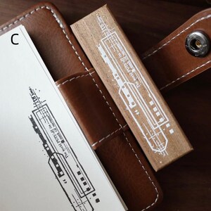 May include: A rectangular wooden rubber stamp featuring a white graphic print of a train and the text The world PUTS off its mask of vastness to its lovers. The stamp is positioned on a brown leather notebook cover with white stitching.