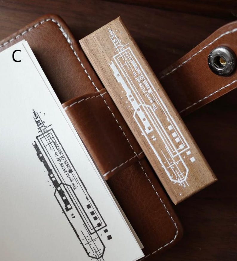May include: A rectangular wooden rubber stamp featuring a white graphic print of a train and the text The world PUTS off its mask of vastness to its lovers. The stamp is positioned on a brown leather notebook cover with white stitching.