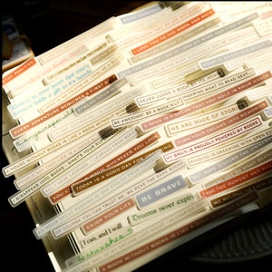 May include: A collection of colorful, rectangular stickers with inspirational quotes about books and reading. The stickers feature a variety of fonts and colors, including brown, white, blue, and gold. The text on the stickers includes phrases like "Stay Curious", "Every Adventure Requires a First Step", "We Are Made of Stories", and "Be Brave".