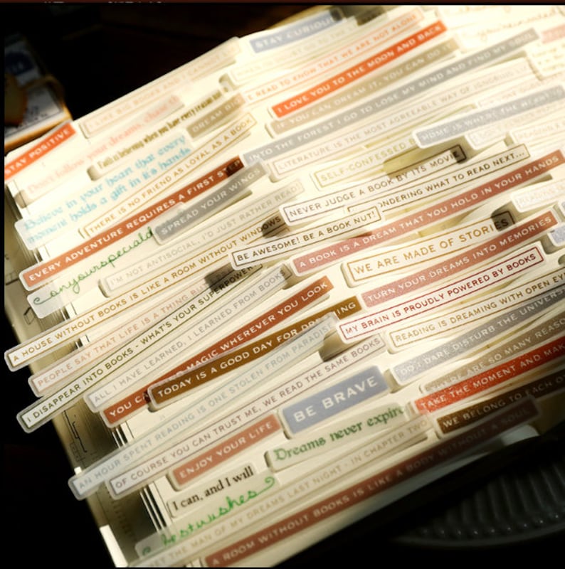 May include: A collection of colorful, rectangular stickers with inspirational quotes about books and reading. The stickers feature a variety of fonts and colors, including brown, white, blue, and gold. The text on the stickers includes phrases like "Stay Curious", "Every Adventure Requires a First Step", "We Are Made of Stories", and "Be Brave".