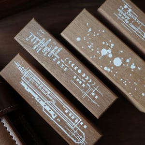May include: Three rectangular wooden blocks featuring white printed designs. One block displays a moon phase cycle and text, another shows a paint splatter pattern, and the third features a linear graphic with the text, The world PUTS off its mask of vastness.
