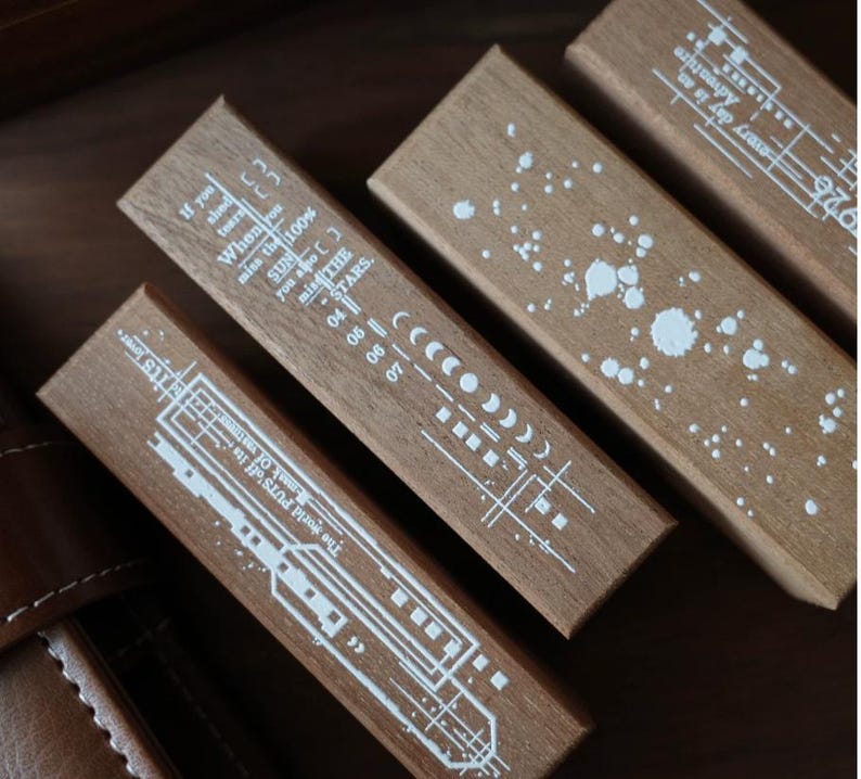 May include: Three rectangular wooden blocks featuring white printed designs. One block displays a moon phase cycle and text, another shows a paint splatter pattern, and the third features a linear graphic with the text, The world PUTS off its mask of vastness.