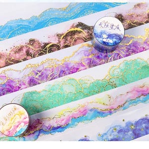 May include: Decorative washi tape rolls and strips with watercolor designs in shades of blue, purple, pink, and green, accented with gold foil. The tape rolls feature Chinese characters and colorful abstract art.