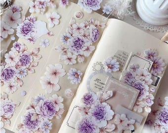 Puede incluir: Una hoja de pegatinas transparentes con diseños florales de color morado y blanco. Las pegatinas presentan flores y hojas delicadas, perfectas para agregar un toque de elegancia a diarios, planificadores o proyectos de manualidades.