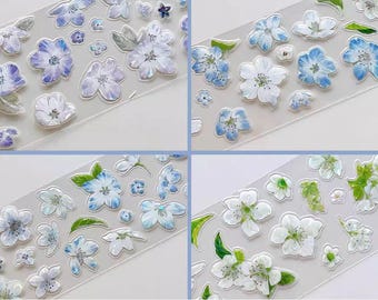 Puede incluir: Hojas de pegatinas transparentes con diseños florales. Las pegatinas muestran flores blancas, azules y moradas con hojas verdes. Las flores tienen un acabado brillante y están en varios tamaños y formas.