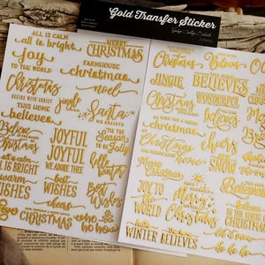 May include: Two white sheets of gold transfer stickers featuring various Christmas-themed phrases and words in cursive and block fonts. The header card reads Gold Transfer Sticker. Phrases include Merry Christmas, Joy to the World, and Believe.
