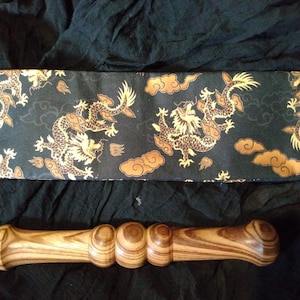 Può includere: Un rullo da massaggio in legno con un motivo a strisce marrone e marrone chiaro. Il rullo ha un'estremità arrotondata e una serie di protuberanze lungo la sua lunghezza. Il rullo è appoggiato su un tessuto nero con un motivo di drago dorato e nero.