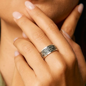 May include: A silver ring with a detailed, intricate design. The ring is worn on the finger of a person with light skin.