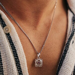 May include: A silver chain necklace with a silver pendant featuring a pink gemstone in the center. The pendant is a geometric design with a square shape and intricate details.