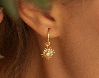May include: A gold hoop earring featuring a sun-shaped charm with a central iridescent stone. The charm has radiating rays extending from the center, creating a sunburst design. The earring is suspended from a small, thin gold hoop.