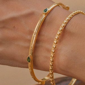 Può includere: Due braccialetti d'oro su un polso. Uno ha un design curvo con due pietre verdi a forma di goccia. L'altro braccialetto è ricoperto da piccole perle d'oro in rilievo. I gioielli sono esposti su una carnagione chiara.
