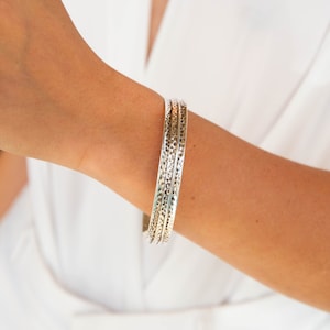 May include: A set of three silver bangles with a hammered texture. The bangles are stacked on a person's wrist.