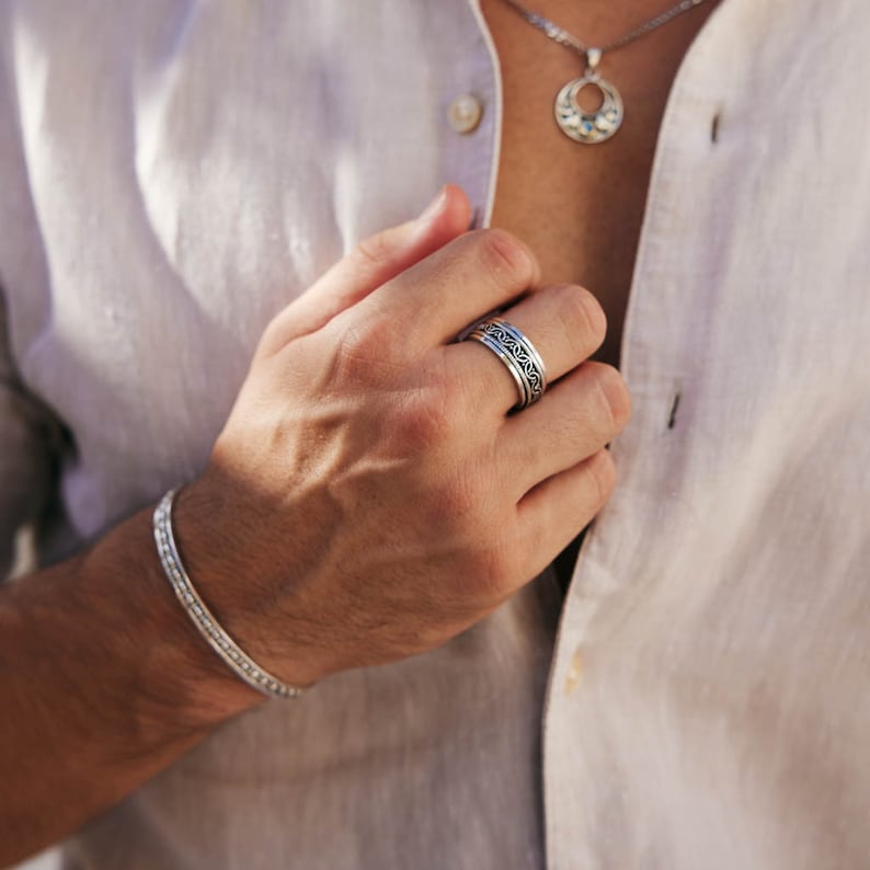 May include: A silver ring with intricate designs. The ring is worn on the index finger of a person's hand. The person is wearing a white linen shirt.