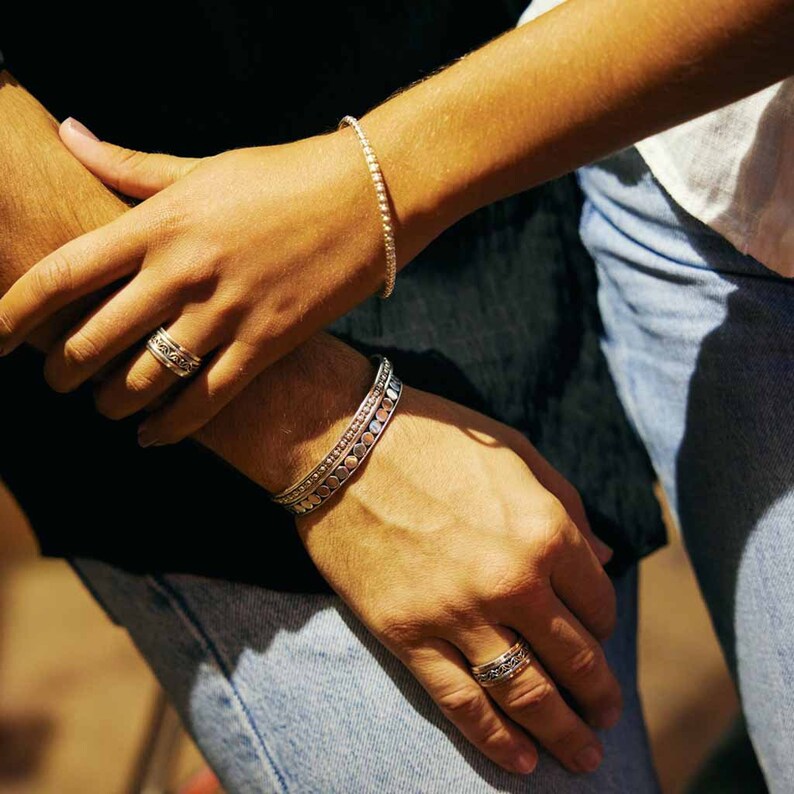 May include: Two silver bracelets and two silver rings on a person's wrists and fingers. The bracelets are both silver and have a textured design. The rings are both silver and have a simple design.