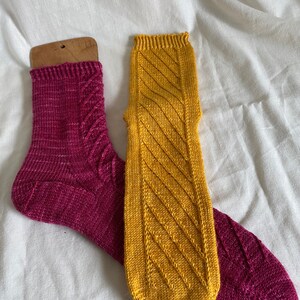 May include: A pair of hand-knitted socks, one in a deep pink color and the other in a bright yellow color. Both socks feature a diagonal cable knit pattern.