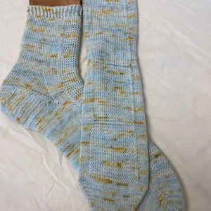 May include: A pair of hand-knitted socks in shades of light blue, yellow, and white. The socks are made of a soft, textured yarn and are displayed on a white surface. The socks have a ribbed cuff and a patterned design.