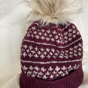 May include: A burgundy knit hat with a white geometric pattern and a faux fur pom-pom. The hat has a ribbed brim and a soft, textured appearance. The pom-pom is a light beige color.
