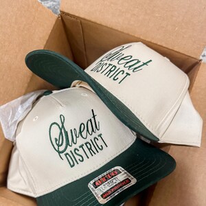 May include: Two beige baseball caps with green brims. The caps have the text "Sweat District" embroidered on the front in green cursive. One cap has a label that reads "O'THIGGS Classic Fit 6 7/8 MID PROFILE MAXIMUM COMFORT"