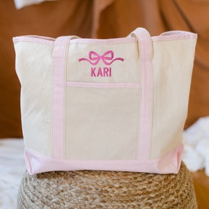 May include: A beige canvas tote bag with pink trim and handles. The bag has a pink embroidered bow and the name "KARI" on the front.