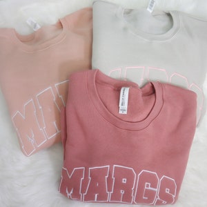 May include: Three sweatshirts with the name "MARCS" written in white on each. The sweatshirts are in pink, light pink, and gray.