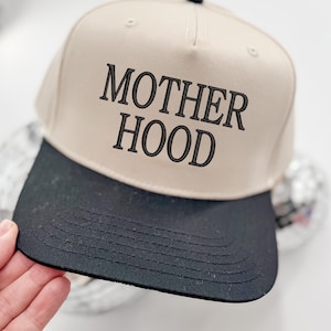 May include: A beige and black baseball cap with the text "MOTHERHOOD" embroidered in black on the front.