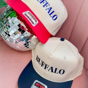 May include: Two beige baseball caps with black and red brims. The front of each cap has the word "BUFFALO" embroidered in blue and black lettering. The cap on the left has a red brim and the cap on the right has a black brim.