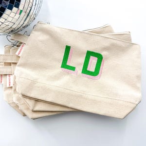 May include: Stack of three beige canvas zippered pouches with green and pink embroidered initials "LD" on the front. The pouches are labeled "CB STATION" in red.