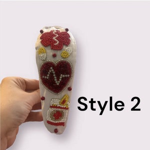 May include: White headband with red, yellow, and white beaded designs. The designs include a heart with a heartbeat symbol, a pill bottle, and a medical symbol. The headband is labeled "Style 2".