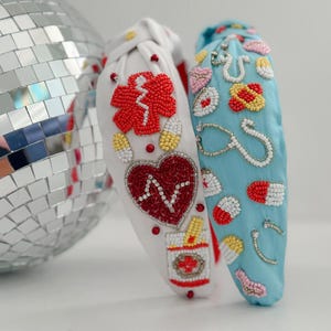 May include: Two beaded headbands with a medical theme. One headband is white with red, white, and blue beads, and the other is blue with white, red, and yellow beads. The beads are arranged to form medical symbols like a stethoscope, pills, and a heart.