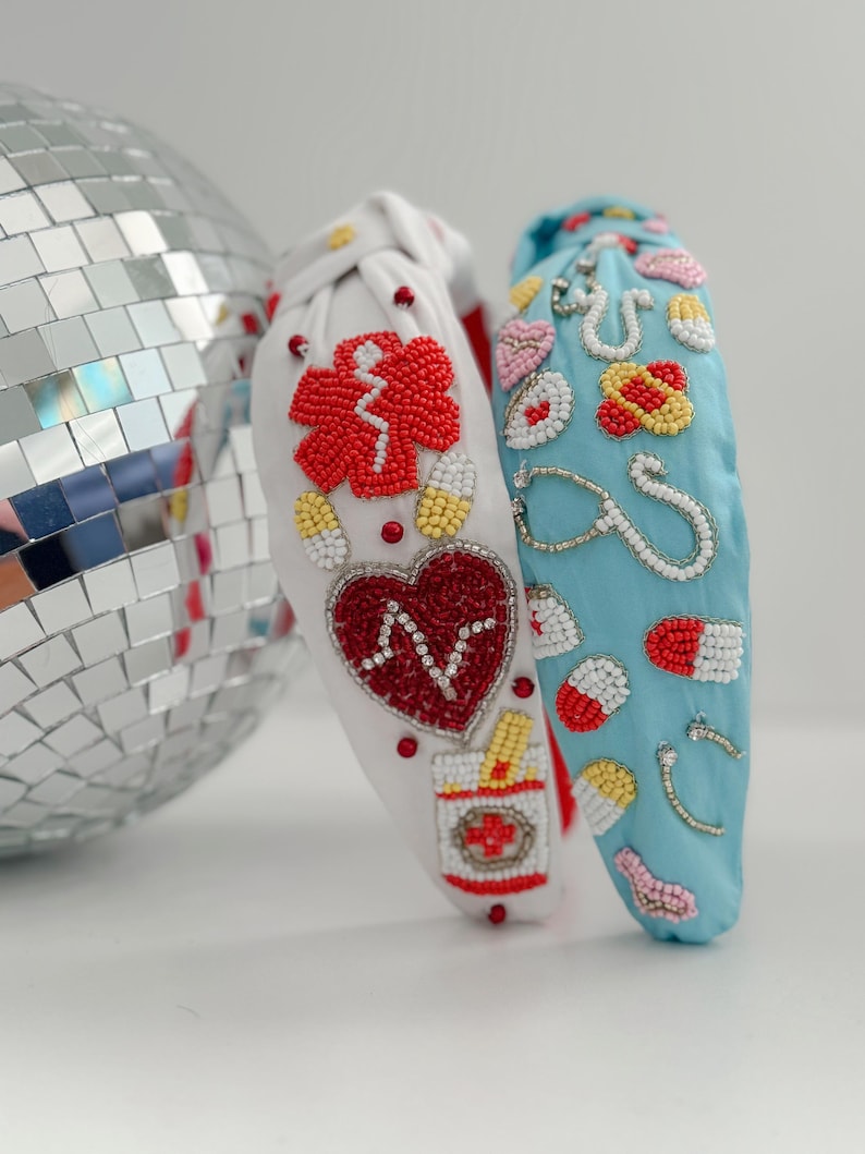 May include: Two beaded headbands with a medical theme. One headband is white with red, white, and blue beads, and the other is blue with white, red, and yellow beads. The beads are arranged to form medical symbols like a stethoscope, pills, and a heart.