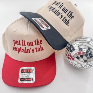May include: Two beige baseball caps with black and red brims. The caps have the text "put it on the captain's tab." embroidered in red on the front. One cap has a black brim and the other has a red brim. The caps are sitting on a white surface.