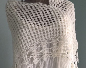 May include: A white crochet shawl draped over a white dress. The shawl features a geometric openwork design and a scalloped edge. The dress has floral embroidery and tassels. The overall style is bohemian and feminine.
