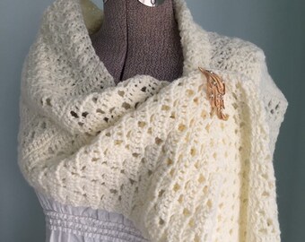 May include: A cream-colored, crocheted shawl draped over a dress form. The shawl features an open, lacy design and is secured with a gold-toned brooch. The dress form is wearing a white dress. The background is a solid light blue.