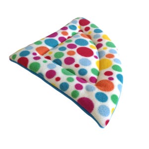 May include: A triangular pet bed featuring a white base with a colourful polka dot pattern in shades of red, blue, green, orange, and yellow. The bed has a raised, padded border and a soft, plush texture with a solid blue fabric trim along the edge.